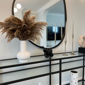 May include: A black metal console table with glass shelves. The table has a white ceramic vase with dried pampas grass, a black knotted sculpture, and a white ceramic vase with gold dots.