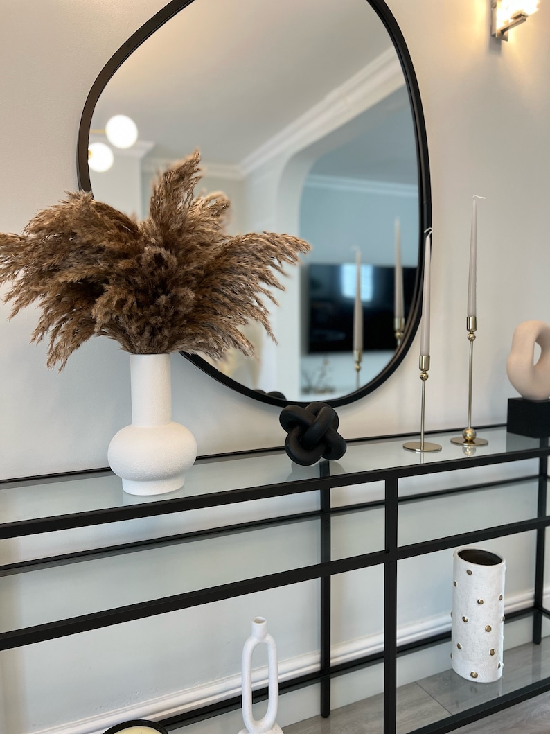 May include: A black metal console table with glass shelves. The table has a white ceramic vase with dried pampas grass, a black knotted sculpture, and a white ceramic vase with gold dots.