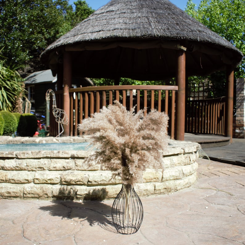 May include: A dried pampas grass bouquet in a black wire vase. The vase is sitting on a stone wall in front of a wooden gazebo.