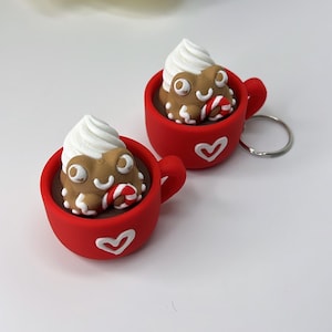 May include: Two red coffee cup keyrings with white heart details. Each cup holds a brown creature with white whipped cream and a candy cane. The keyrings are on a white surface with floral accents.