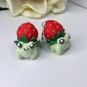 May include: Two small, light green frog figurines with white bellies and dark brown eyes, each topped with a red strawberry featuring yellow seeds and green leaves. One frog is a standalone figurine, while the other is attached to a metal keychain ring.