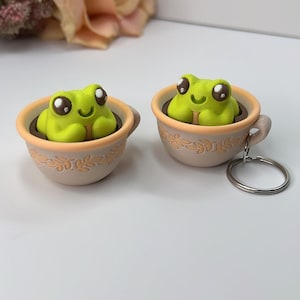 May include: Two lime green frog figurines with large eyes sit inside small, beige teacups. The teacups have a light orange rim and a decorative leaf pattern. One teacup has a silver keychain attached. The background includes a blurred peach-colored flower.