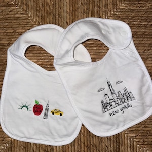 May include: Two white baby bibs with embroidered New York City designs. One bib features a red apple, a yellow taxi, and a cityscape. The other bib displays the New York City skyline with the words "new york".