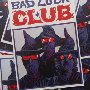 Op de afbeelding: Een witte kaart met de tekst "Bad Luck Club" in rode en blauwe letters. De kaart toont een illustratie van drie mensen die zwarte hoeden en rode zonnebrillen dragen.