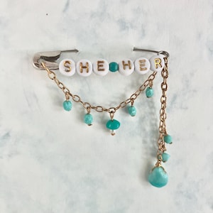 May include: A handmade safety pin with the word "SHE HER" spelled out in white and gold beads. The pin features a gold chain with turquoise beads and a turquoise mushroom charm, creating a unique and personalized accessory.
