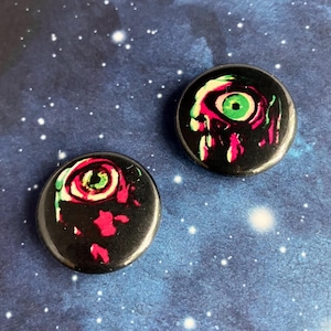 May include: Two black button pins with a graphic of a pink and green eye on a black background.