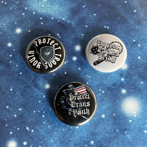 May include: Three round buttons with the text "Protect Trans Youth" in various designs. One button is black with a shield and sword design. Another is white with a spiked club design. The third is black with a figure holding a trans pride flag.