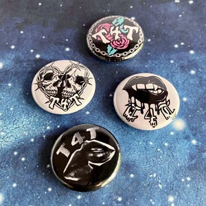 May include: Four black and white button pins with different designs. The first pin features a heart with two skulls inside and the text "TAT" below. The second pin features a rose and the text "TAT" above. The third pin features a dripping mouth with fangs and the text "TAT" below. The fourth pin features a hand with the text "TAT" above.