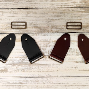May include: A selection of guitar straps and hardware. Two black and two brown leather guitar straps are shown with metal hardware. Two metal rectangle sliders are also included. The straps have a hole at the top for attachment.