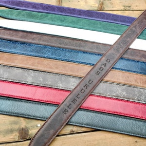 May include: A brown leather guitar strap with the word "DAD" embossed on it. The strap is lying on top of a stack of other guitar straps in various colors.