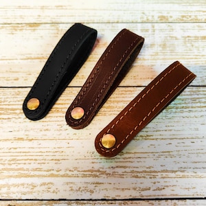 以下が含まれることがあります： 黒、濃い茶色、明るい茶色の3つの革製引き出しプル。各プルは、コントラストのある糸でステッチされ、真鍮色のリベットで固定されています。プルは明るい色の木製の表面に配置されています。