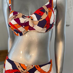 May include: A white, orange, red, and purple patterned bikini top and bottom. The top has a bow in the center and the bottom is a high-waisted, short style.