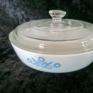 May include: A round white casserole dish with a clear glass lid and a knob handle. The exterior of the white bowl features a blue floral pattern consisting of three stylized flowers with leaves centered on the side of the cooking vessel.