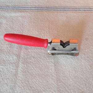 May include: A knife sharpener with a red wooden handle and a silver-colored metal sharpening mechanism. The sharpener has two orange-colored sharpening wheels. The handle is attached to the sharpening mechanism with a metal piece.