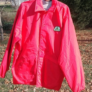 May include: A bright red nylon jacket with a green and white logo on the left chest. The jacket has a snap closure and a drawstring hood.