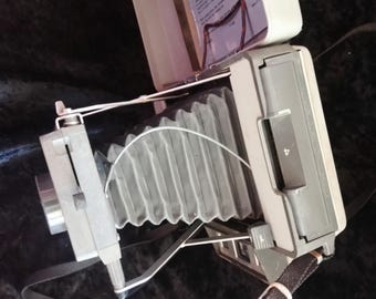 Puede incluir: Una cámara Polaroid vintage de color gris y blanco. La cámara tiene un sistema de fuelle y una correa. La cámara tiene el número "4" en el lateral.
