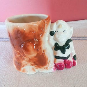 May include: A ceramic planter shaped like a tree stump with a whimsical gnome figure leaning against it. The gnome is painted in white, green, and pink with a black eye.