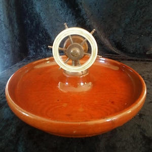 May include: A decorative wooden bowl with a ship's wheel centerpiece. The bowl is a rich, reddish-brown color with a glossy finish. The ship's wheel is made of metal and a white material, adding a nautical touch to the piece.