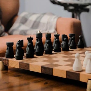 May include: A chess set featuring matte black pieces with metallic accents and a wooden board with alternating light and dark square tiles. The pieces have a tapered, geometric design, and the board is elevated on small metallic feet.