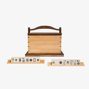 May include: A wooden box with a hinged lid and a handle, used for storing mahjong tiles. The box is made of light and dark wood and has a natural finish. Two sets of mahjong tiles are displayed on wooden racks.