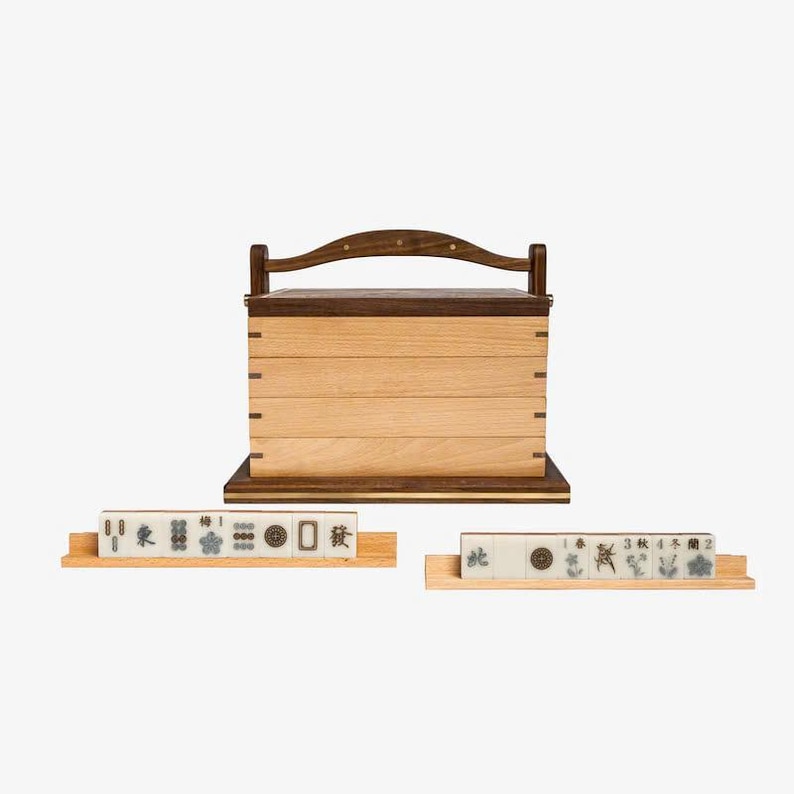May include: A wooden box with a hinged lid and a handle, used for storing mahjong tiles. The box is made of light and dark wood and has a natural finish. Two sets of mahjong tiles are displayed on wooden racks.