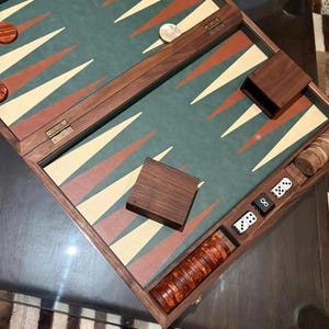 May include: A wooden backgammon set with a green playing surface, featuring red, beige, and white triangular points. The set includes wooden game pieces, dice, and a wooden dice cup. The set is displayed on a glass table.