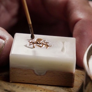 May include: A close-up of a small, white, rectangular box with a gold design on top. A wooden stick with a brush on the end is being used to apply gold paint to the design.