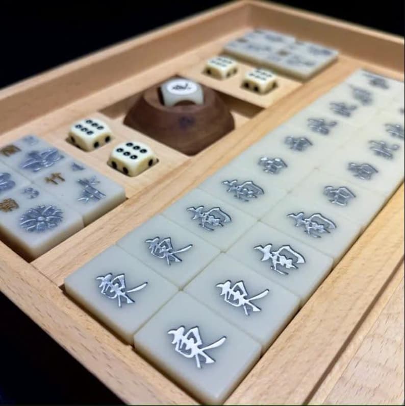 May include: A wooden box containing a set of mahjong tiles. The tiles are white with black and silver characters. There are also four dice and a wooden stand with a single die on top.