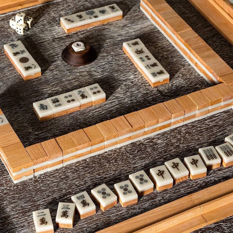 May include: A wooden mahjong set with white tiles featuring Chinese characters and symbols. The tiles are arranged on a wooden board with a dark brown surface. The set includes dice and a wooden stand.