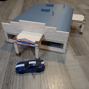 Peut inclure: Une voiture jouet bleue et blanche est placée devant un bâtiment miniature avec une enseigne indiquant "Marshall Performance Motors". Le bâtiment est fait de carton mousse blanc avec un toit bleu et une texture de pierre.