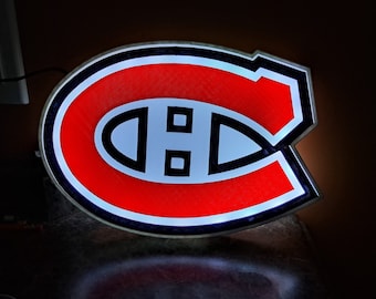 May include: A red, white, and blue illuminated sign with the Montreal Canadiens logo. The logo is a stylized "C" with a white center and black outline.