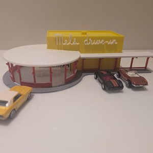 May include: A miniature yellow Mel's Drive-In restaurant model with a white roof and red accents. Three toy cars are parked nearby, including a yellow car with a white roof, and two other cars. The sign on the building reads "Mels drive-in".