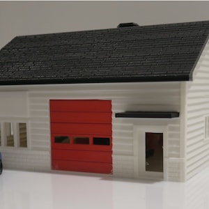 May include: A white model house with a black roof, red garage door, and a small blue car in front. The house has a small window on the side and a door on the front.