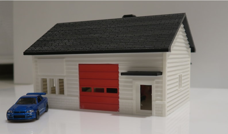 May include: A white model house with a black roof, red garage door, and a small blue car in front. The house has a small window on the side and a door on the front.