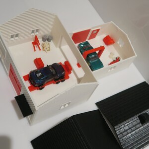 May include: A white toy garage with red doors and a blue car outside. Inside the garage are two toy cars, one blue and one green, on a red lift. There are also various tools and accessories inside the garage.