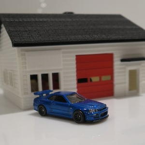 May include: A blue toy car parked in front of a white toy house with a red garage door.