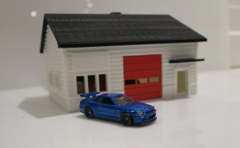 May include: A blue toy car parked in front of a white toy house with a red garage door.
