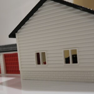 May include: A white model house with a red garage door and two windows. The house has a black roof and a brick base.