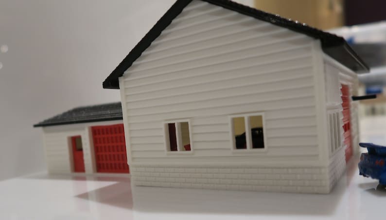 May include: A white model house with a red garage door and two windows. The house has a black roof and a brick base.