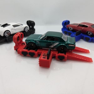 Könnte beinhalten: Ein roter Spielzeug-Autotransporter mit zwei Rampen und einem grünen Spielzeugauto oben drauf. Der Transporter hat schwarze Räder und ein schwarz-rotes Design. Im Hintergrund befinden sich zwei weitere Spielzeugautos, eines weiß und eines rot.