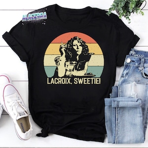 May include: A black t-shirt featuring a vintage-style illustration of a woman holding a drink and smoking a cigarette. The illustration is set against a sunset background with a rainbow of colours. The text "LACROIX, SWEETIE!" is printed below the illustration.
