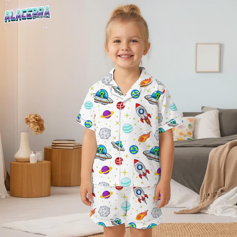 Puede incluir: Conjunto de pijama blanco con un colorido estampado de tem&aacute;tica espacial con cohetes, planetas, ovnis y extraterrestres. La parte superior de manga corta y los pantalones cortos tienen botones y ribetes blancos. El pijama lo lleva un ni&ntilde;o peque&ntilde;o.