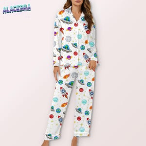 Puede incluir: Conjunto de pijama blanco con un colorido estampado de tem&aacute;tica espacial. El conjunto incluye una camisa con botones y pantalones a juego, con cohetes, planetas, ovnis y estrellas. El pijama tiene un corte holgado y est&aacute; hecho de un material suave.
