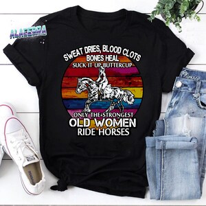 Op de afbeelding: Zwart T-shirt met een vintage zonsondergang-afbeelding en de tekst "Sweat dries, blood clots, bones heal, suck it up buttercup. Only the strongest old women ride horses."