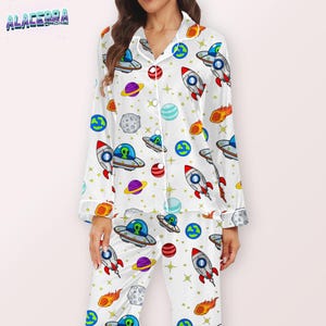 Puede incluir: Conjunto de pijama blanco con un estampado de tem&aacute;tica espacial. El dise&ntilde;o incluye cohetes, planetas, ovnis y estrellas. El pijama tiene una camisa abotonada con cuello y pantalones a juego. La marca "ALACERRA" es visible.