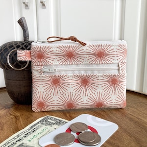 May include: A small white zippered pouch with a red and white geometric pattern. The pouch has a brown leather strap and a silver zipper pull. The pouch is sitting on a wooden surface with a dollar bill and some coins.