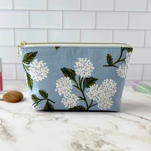 May include: A light blue cosmetic bag with a floral pattern of white flowers and green leaves. The bag has a gold zipper and is sitting on a white surface. Makeup brushes and tubes are in the background.