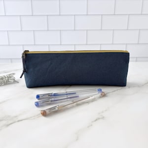 May include: A navy blue pencil case with a gold zipper, next to three pens and a pair of white earbuds. The pencil case is rectangular and made of a sturdy fabric. The pens are clear with blue and brown ink.
