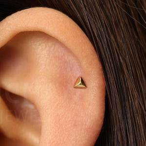 Peut inclure: Gros plan sur une oreille avec une petite boucle d'oreille triangulaire dorée. La boucle d'oreille est positionnée sur le cartilage supérieur de l'oreille. Le teint est clair, et la boucle d'oreille est le point central.