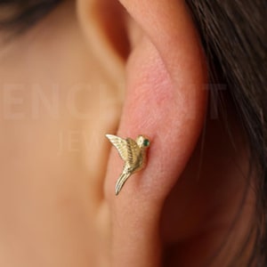 May include: A gold-coloured hummingbird stud earring with a small green gem eye. The earring is worn on the ear of a person with dark hair. The earring is a small, detailed sculpture of a bird.